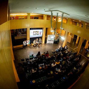 Blick von oben auf eine Veranstaltung mit zwei Personen an einem Tisch vor einer Leinwand und einem Publikum in einem modernen, holzverkleideten Raum.