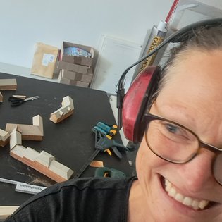 Nahaufnahme einer lächelnden Frau mit Brille und Gehörschutz vor einem Tisch mit Holzbauteilen und Werkzeugen