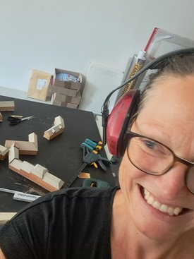 Nahaufnahme einer lächelnden Frau mit Brille und Gehörschutz vor einem Tisch mit Holzbauteilen und Werkzeugen