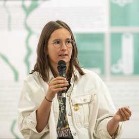 Lucia Parbel hält ein Mikrofon in der Hand. Sie hat braunes Haar, das ihr bis über die Schulter fällt und trägt eine Brille.
