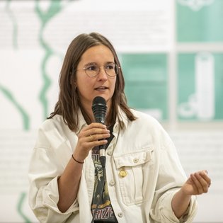 Lucia Parbel hält ein Mikrofon in der Hand. Sie hat braunes Haar, das ihr bis über die Schulter fällt und trägt eine Brille.