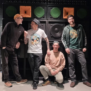 Vier Männer vor einer großen Lautsprecherwand, einer trägt ein weißes T-Shirt mit Aufdruck, einer eine grüne Jacke mit Streifen an den Ärmeln, einer eine schwarze Jacke und einer einen hellen Pullover und sitzt hockend