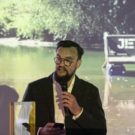 Marc Gegenfurtner, Amtsleiter des Kulturamts Stuttgart, während seiner Eröffnungsrede. Er hält ein Mikrofon in der einen Hand und in der anderen ein Smartphone. Im Hintergrund auf einer Leinwand: ein Foto des Festival-Floßes auf einem Gewässer.