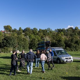 Mehrere Personen bilden einen Kreis. Ein schwarzes Band, von den Personen am Rücken festgehalten, begrenzt den Kreis. Im Hintergrund steht ein größeres Auto mit einem Anhänger, der mit Kanus vollgeladen ist.