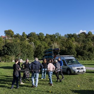Mehrere Personen bilden einen Kreis. Ein schwarzes Band, von den Personen am Rücken festgehalten, begrenzt den Kreis. Im Hintergrund steht ein größeres Auto mit einem Anhänger, der mit Kanus vollgeladen ist.