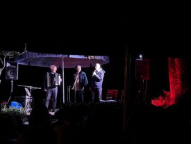 Drei Musiker auf einer kleinen Außenbühne bei Nacht: links spielt ein Mann Akkordeon, daneben singen zwei Männer ins Mikrofon. Vor der Bühne sitzen Zuschauer im Dunkeln.