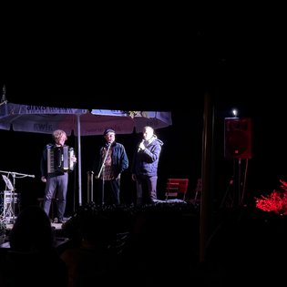 Drei Musiker auf einer kleinen Außenbühne bei Nacht: links spielt ein Mann Akkordeon, daneben singen zwei Männer ins Mikrofon. Vor der Bühne sitzen Zuschauer im Dunkeln.