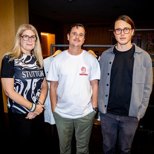 Drei Personen stehen nebeneinander, Nina Preuss links trägt ein schwarz-weißes Shirt mit dem Schriftzug 'STUTTGART', Ingmar Volkmann mittig trägt ein weißes T-Shirt mit kleinem rotem Logo, Nils Unterharnscheidt rechts trägt eine graue Jacke über schwarzem Shirt.