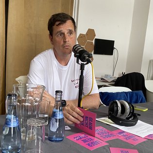 Podcast-Moderator Ingmar Volkmann sitz an einem Tisch und spricht in ein Mikrofon. Vor ihm auf dem Tisch stehen verschiedene Getränke und es liegen pinke Postkarten aus dem Projekt »Schlamassel Tov« aus.