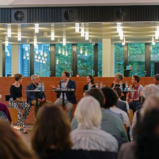 ca 100 Publikumsgäste schauen auf das Podium, das Podium sitzt auf Barhockern vor einer Backsteinmauer, im Hintergrund sieht man die Pflanzen im Innenhof der Mensa