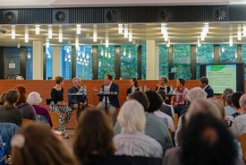 ca 100 Publikumsgäste schauen auf das Podium, das Podium sitzt auf Barhockern vor einer Backsteinmauer, im Hintergrund sieht man die Pflanzen im Innenhof der Mensa