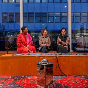 Drei Personen sitzen auf einer Bühne vor einem großen Fenster und führen eine Diskussion, im Vordergrund sind Zuschauer zu sehen.