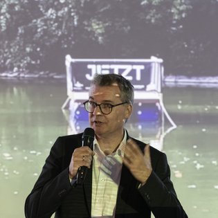 Dr. Alexander Lahl, Regionaldirektor/Verband Region Stuttgart, während seiner Eröffnungsrede. Er hält ein Mikrofon in der Hand und richtet sich mit Gesten ans Publikum. Im Hintergrund auf einer Leinwand: ein Foto des Festival-Floßes auf einem Gewässer.