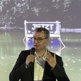 Dr. Alexander Lahl, Regionaldirektor/Verband Region Stuttgart, während seiner Eröffnungsrede. Er hält ein Mikrofon in der Hand und richtet sich mit Gesten ans Publikum. Im Hintergrund auf einer Leinwand: ein Foto des Festival-Floßes auf einem Gewässer.