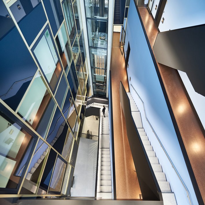 Blick von oben in ein modernes, mehrstöckiges Atrium mit Treppen, gläsernem Aufzugsschacht und Fluren.