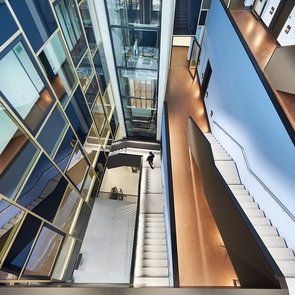 Blick von oben in ein modernes, mehrstöckiges Atrium mit Treppen, gläsernem Aufzugsschacht und Fluren.