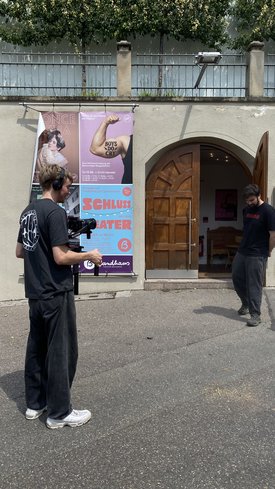 Zwei Männer vor einem Gebäude mit offenem Holztor, einer filmt mit Kamera, an der Wand ein Plakat mit Text und Bildern.