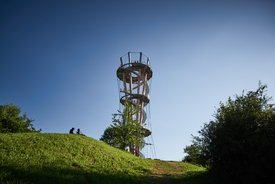 Foto: Blick von unten über eine grüne Wiese hinweg auf den Turm. Auf der Wiese sitzen zwei Personen. Der Himmel ist blau.