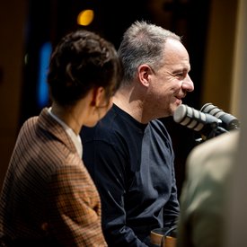 Rocío Alemán und Alexander Zorniger während der Podcast Aufnahme. Sie sitzen beide auf Barhockern und haben Mikrofone vor sich. Der Fokus der Aufnahme liegt auf Alexander Zorniger, Rocío Alemán ist nur von hinten zu sehen.