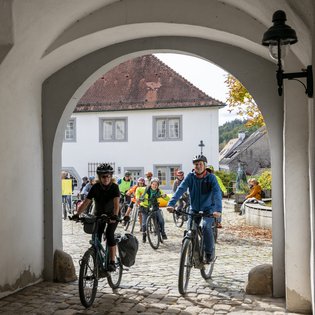 Radfahrer*innen brechen zur Aufführung von Mauricio Kagels Stück »Eine Brise – Flüchtige Aktion für 111 Radfahrer« auf. Sie fahren durch den Torbogen der in den Schlosshof von Schloss Waldenbuch führt.