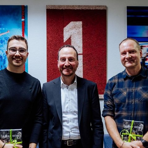 Drei Männer stehen vor einer Wand mit zwei Sportbildern, einer zeigt einen Wasserspringer, der andere eine Curling-Szene, zwei Männer halten Bücher in der Hand.