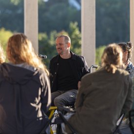 Michael Turinsky auf der Terrasse des Literaturmuseum der Moderne. Turinsky trägt eine dunkle Jacke und ein dunkles Shirt. Er sitzt vor einem Mikrofon und nutzt einen Rollstuhl..
