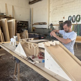 Mann baut in Werkstatt eine Minigolfbahn aus Holz mit Rampe und Ziegenfigur