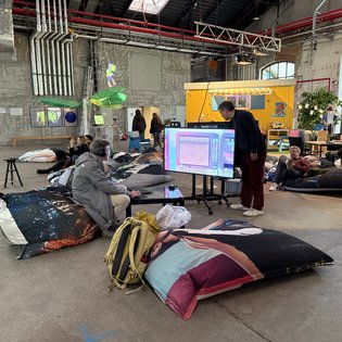 Teilnehmer entspannen auf Sitzsäcken und betrachten digitale Kunst auf einem Bildschirm beim Kunst-im-Game-Symposium.