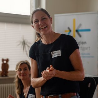 Bettina Pau steht vor einem Banner mit dem Text ‚KulturRegion Stuttgart‘. Im Hintergrund sitzt eine weitere Person mit Namensschild. Auf der Fensterbank befinden sich eine hölzerne Eulenfigur und eine grüne Topfpflanze. Die Szene wirkt wie ein Moment aus einer Veranstaltung oder einer Präsentation in einem Innenraum.