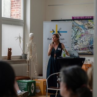 Frau steht an einem Rednerpult vor einem Banner mit der Aufschrift ‚Kultur Region Stuttgart‘. Neben ihr befinden sich ein Skelettmodell sowie eine Topfpflanze auf einem Fenstersims. Auf dem Pult stehen ein Laptop und eine Getränkeflasche, davor mehrere bunte Würfel mit Texten