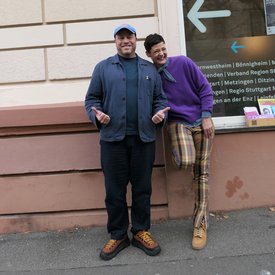 Zwei Personen, Kimsy von Reischach und Jacob Bilabel, lehnen an einer Hauswand, eine trägt blaue Jacke und schwarze Hose, die andere lila Pullover und karierte Hose.