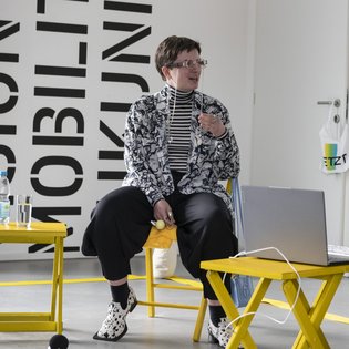 Emilija Tolj sitzt auf einem gelben Stuhl, neben ihr stehen kleine gelbe Tischchen. Sie trägt Kleidung in Schwarz und Weiß und eine Brille mit einer silbernen Brillenkette.