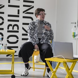 Emilija Tolj sitzt auf einem gelben Stuhl, neben ihr stehen kleine gelbe Tischchen. Sie trägt Kleidung in Schwarz und Weiß und eine Brille mit einer silbernen Brillenkette.