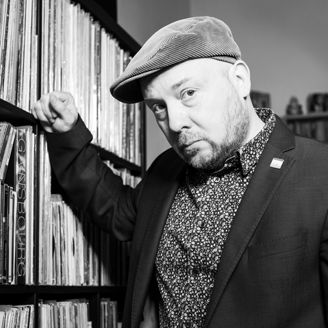 Yuriy Gurzhy lehnt an ein Regal voller Schalplatten. Bartträger Yuriy ist mit eine Kordmütze, ein gemustertes Hemd und ein dunkles Jacket bekleidet. 