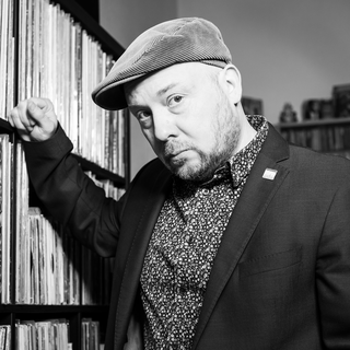 Yuriy Gurzhy lehnt an ein Regal voller Schalplatten. Bartträger Yuriy ist mit eine Kordmütze, ein gemustertes Hemd und ein dunkles Jacket bekleidet. 