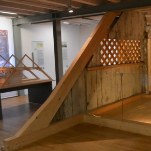 Holzkonstruktion einer historischen Frauenempore aus einer Synagoge, ausgestellt in einem modernen Museumsraum. Die Empore besteht aus gealtertem, weißlich verwittertem Holz mit einer Holzgitteröffnung und wird teilweise von Glaswänden umrahmt. Im Hintergrund befinden sich Informationstafeln und architektonische Modelle.