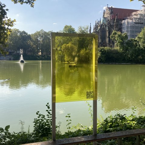 Bild vom Festival Floß auf dem Feuersee in Stuttgart. Fotografiert vom Ufer des Sees durch eine gelbliche Scheibe hindurch. Im Hintergrund die Johanneskirche.