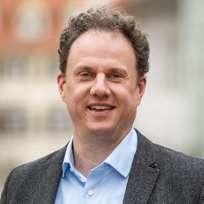 Mann mit lockigem Haar trägt hellblaues Hemd und grauen Sakko steht vor unscharfem Stadt-Hintergrund