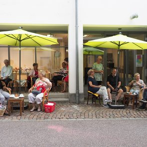 Menschen sitzen unter Sonnenschirmen vor den Café SprechZimmer