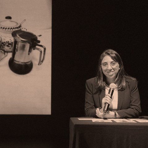 Karina Pino Gallardo sitzt an einem Tisch und hält ein Mikrofon in der Hand. Hinter ihr ist ein Bild mit einem Kaffeekännchen, einer Kaffeetasse, einem Löffel und einer Zuckerdose zu sehen.