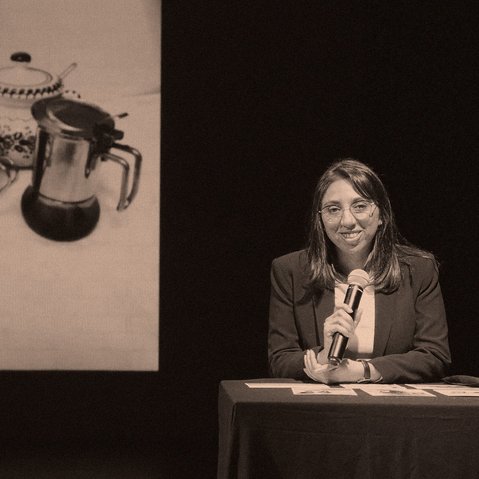Karina Pino Gallardo sitzt an einem Tisch und hält ein Mikrofon in der Hand. Hinter ihr ist ein Bild mit einem Kaffeekännchen, einer Kaffeetasse, einem Löffel und einer Zuckerdose zu sehen.