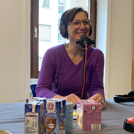 Gäst*in Katja Kranich während der Aufnahme des Podcast. Sie sitzt an einem Tisch, trägt Kopfhörer und hat ein Tischmikrofon vor sich stehen. Auf dem Tisch vor ihr stehen mehrere typische Schuldrinks.
