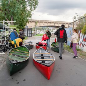 Fünf Kanus liegen auf einem Weg. Ein paar Personen stehen dabei und scheinen sich gerade fürs Kanufahren fertig zu machen oder kommen gerade vom Kanufahren.