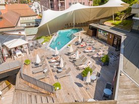 Luftaufnahme einer Holzterrasse mit Pool, Liegestühlen, Sonnenschirmen und mehreren Personen beim Sonnenbaden