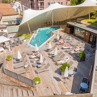 Luftaufnahme einer Holzterrasse mit Pool, Liegestühlen, Sonnenschirmen und mehreren Personen beim Sonnenbaden