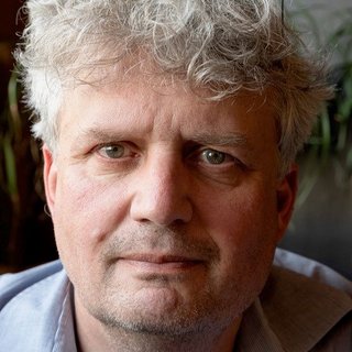 Person mit lockigem, weiß-blondem Haar und blauem Hemd vor unscharfem Hintergrund.