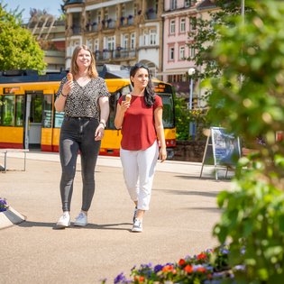 Zwei Frauen gehen in einem Park an Blumenbeeten vorbei, im Hintergrund eine gelbe Straßenbahn und historische Gebäude