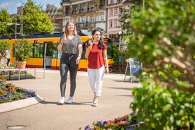 Zwei Frauen gehen in einem Park an Blumenbeeten vorbei, im Hintergrund eine gelbe Straßenbahn und historische Gebäude