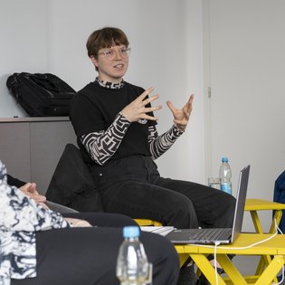 Nisha Toussaint-Teachout sitzt auf einem gelben Stuhl und scheint mit jemandem zu sprechen. Mit den Händen gestikuliert sie. Sie hat kurze, braune Haare und trägt eine Brille.
