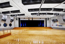 Innenansicht auf den unbestuhlten Kombisaal. Die Wände sind in hellen Tönen gehalten, der Saal verfügt über einen Parkettboden. Hinten im Saal ist eine Bühne zu sehen.